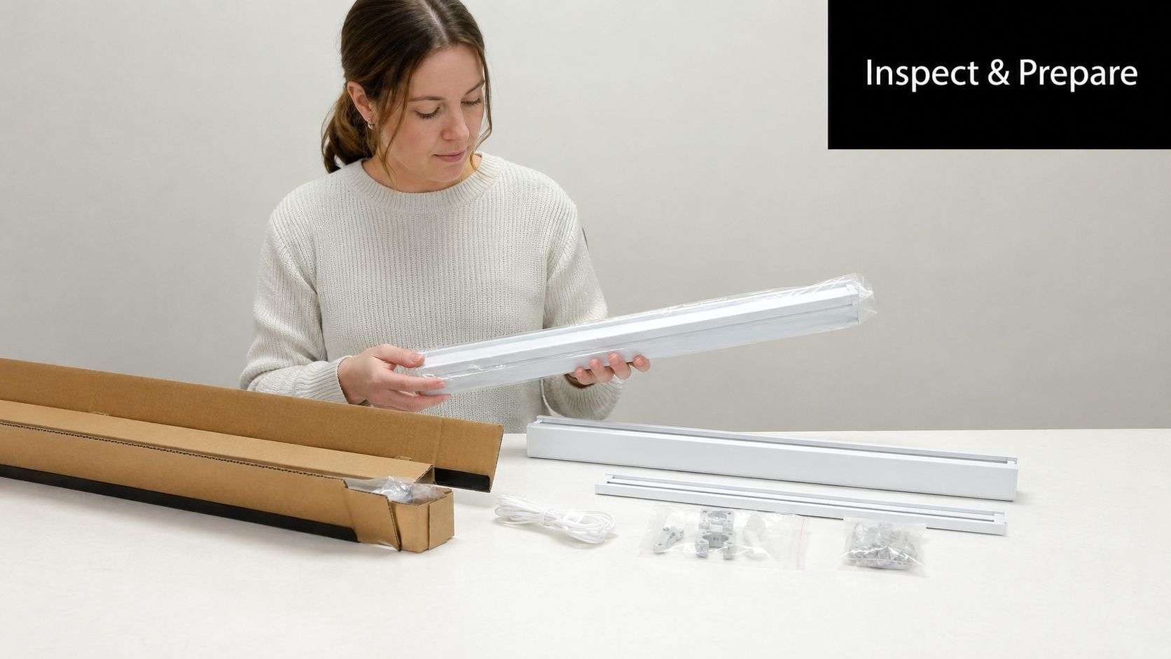 A woman inspecting components for cellular shade installation, including white curtain tracks and mounting hardware on a table.